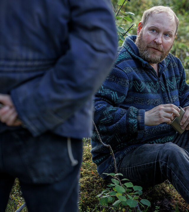 "Wolfssommer – Blutige Spuren - Der Hinterhalt": Thomas (Henrik Dorsin) sitzt im Grünen auf dem Boden. Er hält eine flache Schnapsflasche in den Händen. Ihm gegenüber steht Kat (Eliot Sumner), die nur zum Teil und von hinten zu sehen ist. Sie versteckt ein Messer hinter ihrem Rücken.