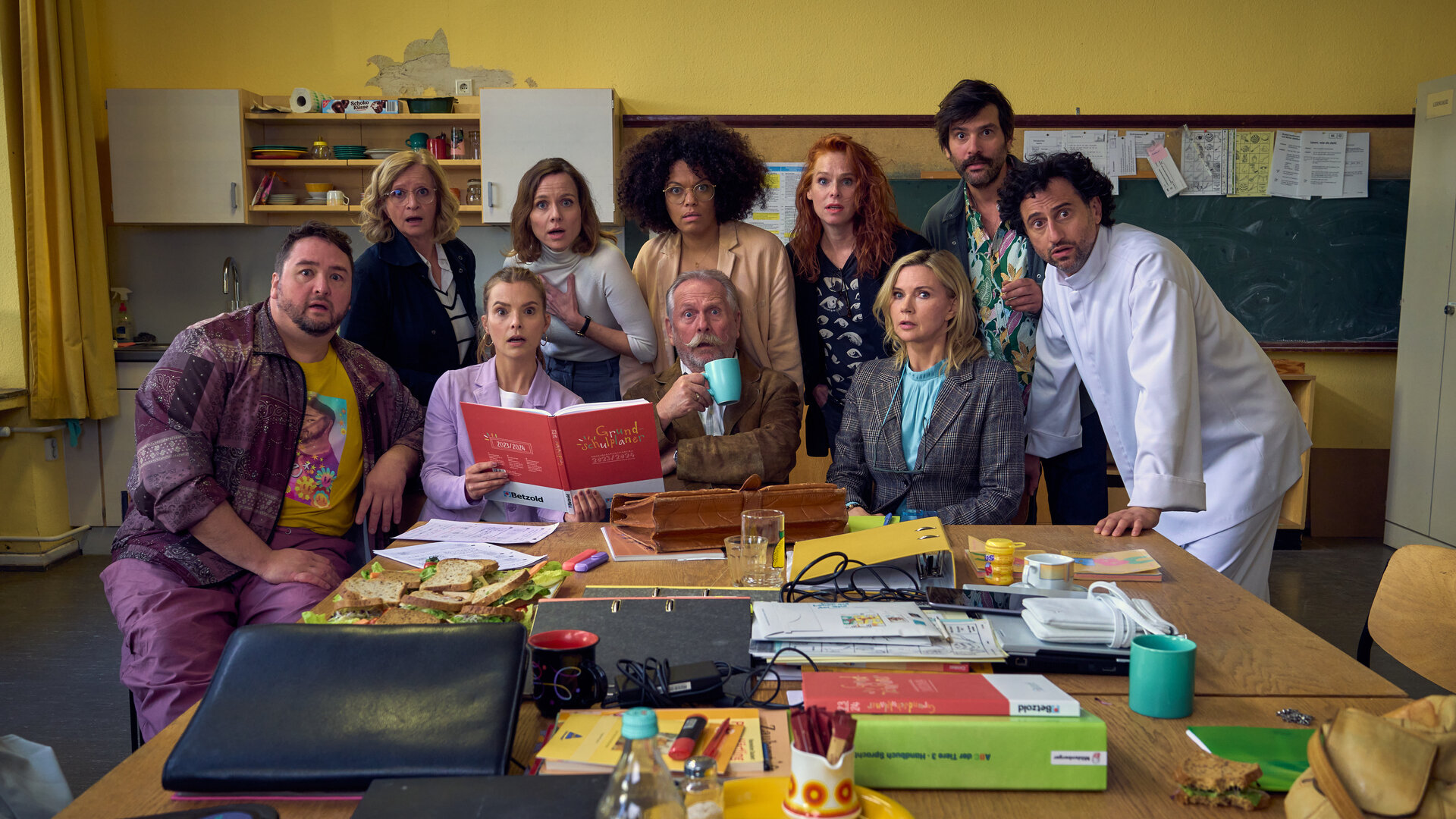 "Andere Eltern - Die 1. Klasse": Malte (Daniel Zillmann), Ini (Johanna Gastdorf), Paula (Maike Jüttendonk), Anita (Nadja Becker), Lehrer (Henning Krautmacher),  Yaa (Rebecca Lina Wilson-Kamm), Nike (Henny Reents), Frau Marx (Veronica Ferres), Jannos (Jasin Challah) und Björn (Serkan Kaya) sitzen gemeinsam im Lehrerzimmer an einem Tisch voller Schulunterlagen und schauen erschrocken in die Kamera.