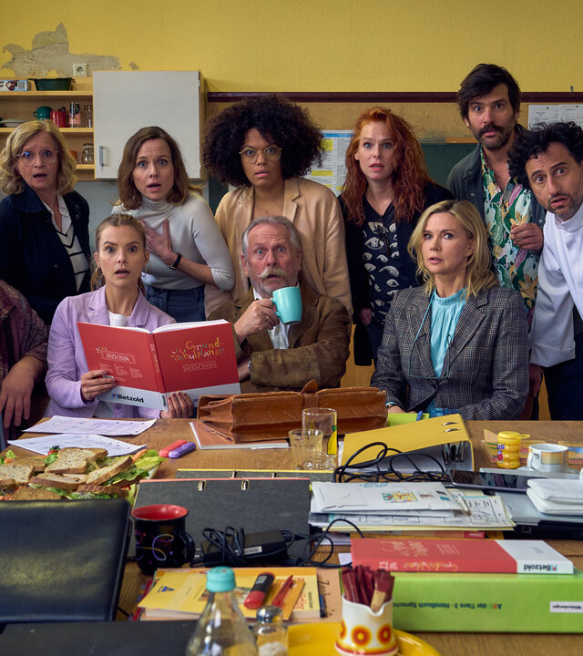 "Andere Eltern - Die 1. Klasse": Malte (Daniel Zillmann), Ini (Johanna Gastdorf), Paula (Maike Jüttendonk), Anita (Nadja Becker), Lehrer (Henning Krautmacher),  Yaa (Rebecca Lina Wilson-Kamm), Nike (Henny Reents), Frau Marx (Veronica Ferres), Jannos (Jasin Challah) und Björn (Serkan Kaya) sitzen gemeinsam im Lehrerzimmer an einem Tisch voller Schulunterlagen und schauen erschrocken in die Kamera.