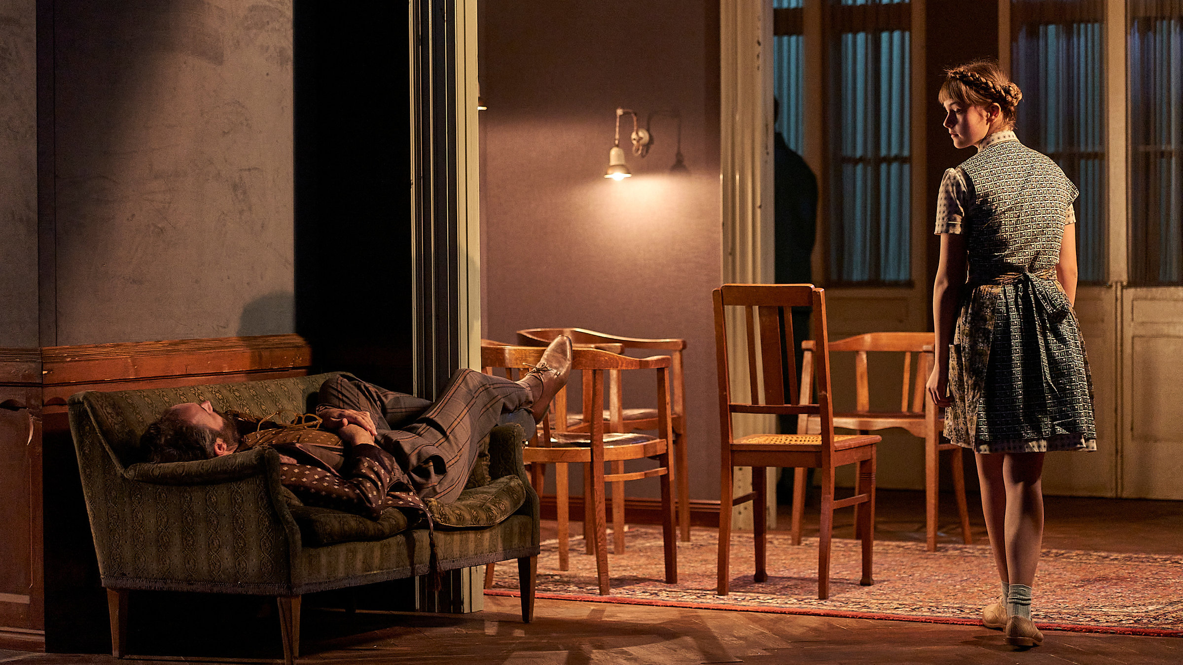 "Starke Stücke: Kinder der Sonne": Das Esszimmer einer großzügigen Altbauwohnung am Abend. Links liegt auf einem kleinen abgenutzten Sofa ein Mann mit den Beinen Richtung Bühnenrückwand. Rechts blickt ein junges Dienstmädchen mit Schürze und geflochtenem Haarkranz im Vorbeigehen auf den Mann. Im Hintergrund mehrere braune Holzstühle auf einem Perserteppich, der Tisch fehlt. Die Türrahmen sind weiß, die Wände lila und im unteren Drittel mit dunklem Holz verkleidet.