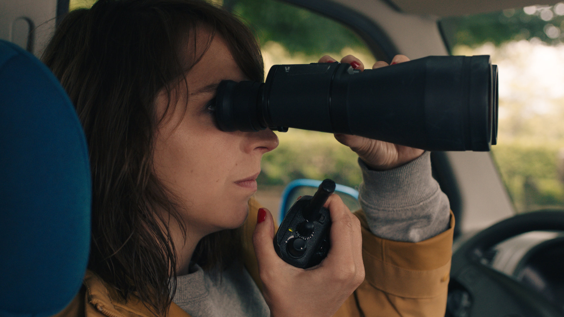 "SOKO Potsdam - Verlust": Luna Kunath (Caroline Erikson) sitzt mit einem Fernglas vor den Augen im Auto. Mit ihrer rechten Hand hält sie sich ein Funkgerät nah an den Mund. Im Hintergrund sieht man durch das Autofenster die grünen Blätter von Bäumen und Büschen.