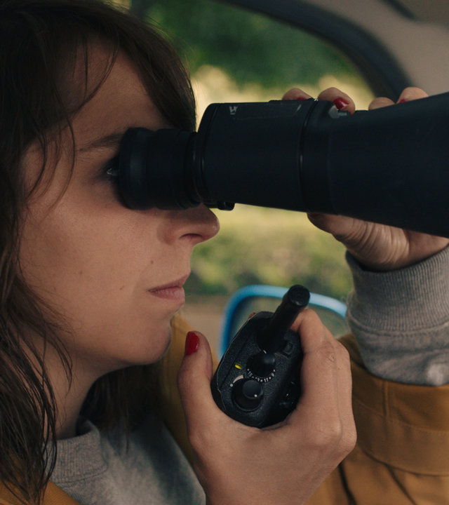"SOKO Potsdam - Verlust": Luna Kunath (Caroline Erikson) sitzt mit einem Fernglas vor den Augen im Auto. Mit ihrer rechten Hand hält sie sich ein Funkgerät nah an den Mund. Im Hintergrund sieht man durch das Autofenster die grünen Blätter von Bäumen und Büschen.