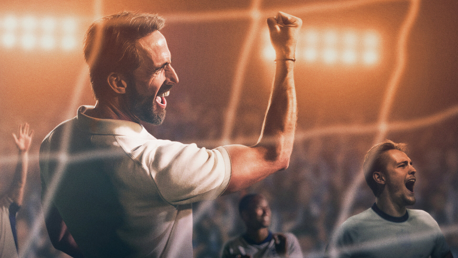 "Dear England (1)": Stilisiertes Bild, im Stadion: Gareth Southgate (Joseph Fiennes) trägt ein weißes Poloshirt und hat den den rechten, muskulösen Arm mit geballter Faust jubelnd in die Höhe gereckt. Hinter ihm befinden sich einige Spieler, dahinter sitzt Publikum auf den Rängen eines Stadions.