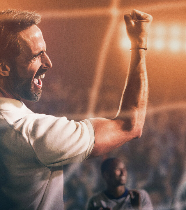 "Dear England (1)": Stilisiertes Bild, im Stadion: Gareth Southgate (Joseph Fiennes) trägt ein weißes Poloshirt und hat den den rechten, muskulösen Arm mit geballter Faust jubelnd in die Höhe gereckt. Hinter ihm befinden sich einige Spieler, dahinter sitzt Publikum auf den Rängen eines Stadions.