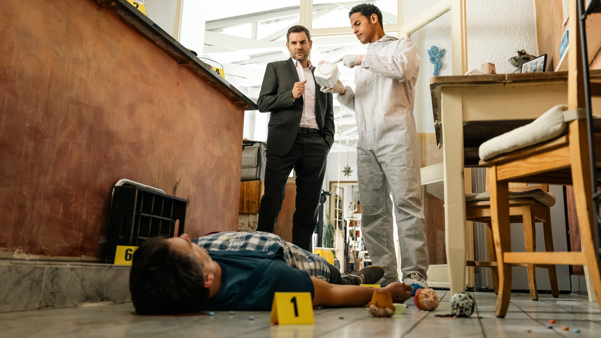 "SOKO Köln - Familie 2.0": Dr. Makena (Stephen Appleton) und Kommissar Jonas Fischer (Lukas Piloty) stehen nebeneinander neben einer hohen, braunen Theke. Dr. Makena trägt einen weißen Schutzanzug und zeigt auf einen weißen Gegenstand, den er Jonas Fischer entgegenhält. Jonas trägt einen dunklen Anzug über einem rosafarbenen Hemd und blickt ernst nach unten zu dem toten David Meyer (Florian Odendahl), der vor ihnen auf dem Rücken am Boden liegt. Aus seinem Kopf fließt Blut auf die Fliesen. Neben ihm liegen drei Muffins und um ihn herum wurden kleine gelben Kärtchen verteilt. An der Seite steht ein Tisch mit zwei Holzstühlen.