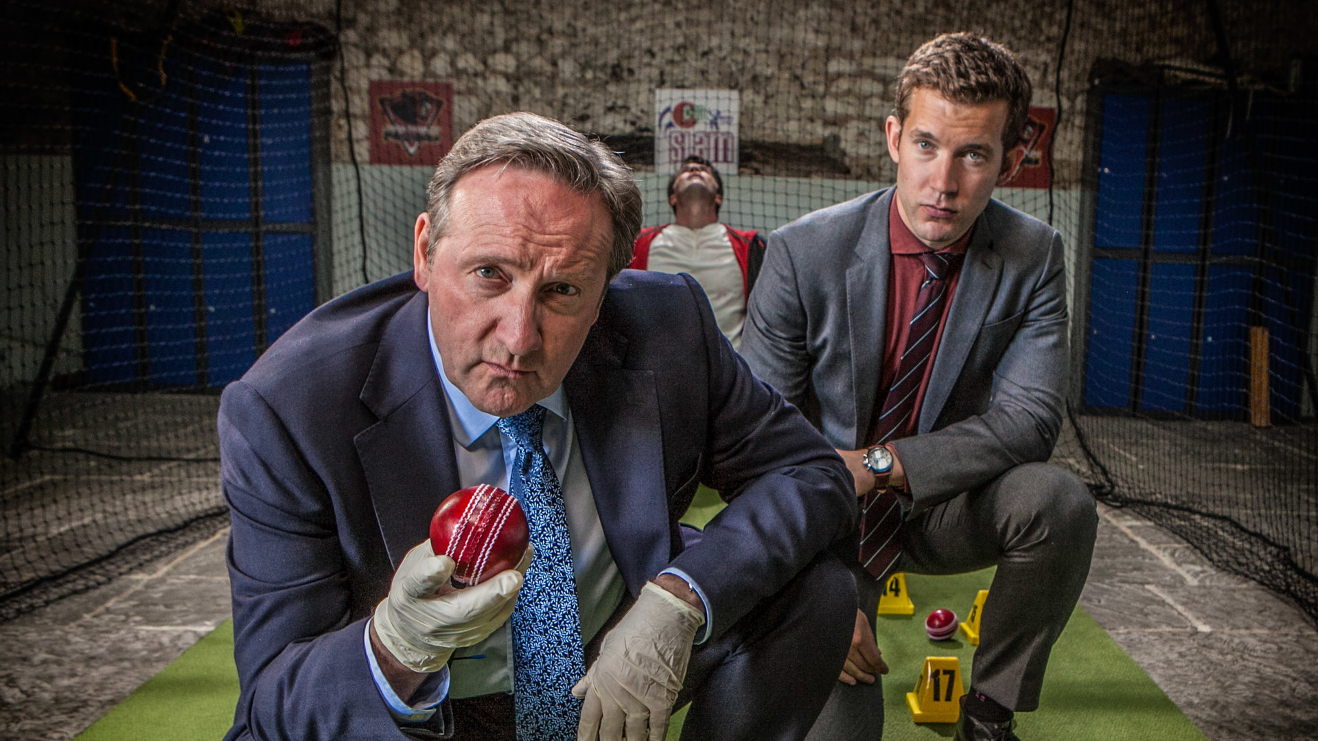 "Inspector Barnaby - Cricket-Fieber": DCI John Barnaby (Neil Dudgeon) und sein Assistent DS Jamie Winter (Nick Hendrix) hocken in einer Cricket-Übungshalle auf einem Stück Kunstrasen, auf dem verstreut ein paar rote Cricket-Bälle liegen. Diese sind mit nummerierten Aufstellern als Tatwaffen gekennzeichnet. Einen der Bälle hält Barnaby in seiner mit Latexhandschuh überstreiften Hand. Im Hintergrund hängt Cricket-Spieler Leo Henderson (Bruce Lawrence) tot im Netz.