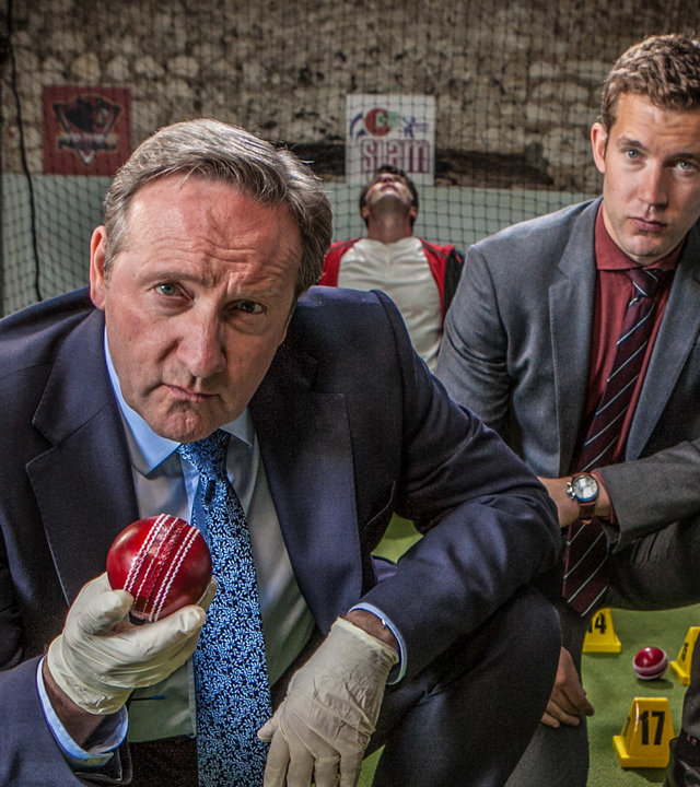 "Inspector Barnaby - Cricket-Fieber": DCI John Barnaby (Neil Dudgeon) und sein Assistent DS Jamie Winter (Nick Hendrix) hocken in einer Cricket-Übungshalle auf einem Stück Kunstrasen, auf dem verstreut ein paar rote Cricket-Bälle liegen. Diese sind mit nummerierten Aufstellern als Tatwaffen gekennzeichnet. Einen der Bälle hält Barnaby in seiner mit Latexhandschuh überstreiften Hand. Im Hintergrund hängt Cricket-Spieler Leo Henderson (Bruce Lawrence) tot im Netz.