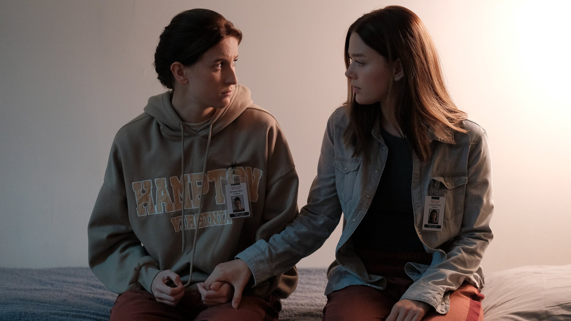 "Fallen - Schuldgefühle": Arriane (Josefine Koenig) und Luce (Jessica Alexander) sitzen nebeneinander auf einem Bett und halten sich an den Händen. Beide blicken sich dabei ruhig und verständnisvoll an.