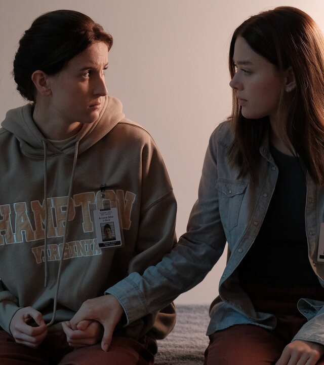 "Fallen - Schuldgefühle": Arriane (Josefine Koenig) und Luce (Jessica Alexander) sitzen nebeneinander auf einem Bett und halten sich an den Händen. Beide blicken sich dabei ruhig und verständnisvoll an.