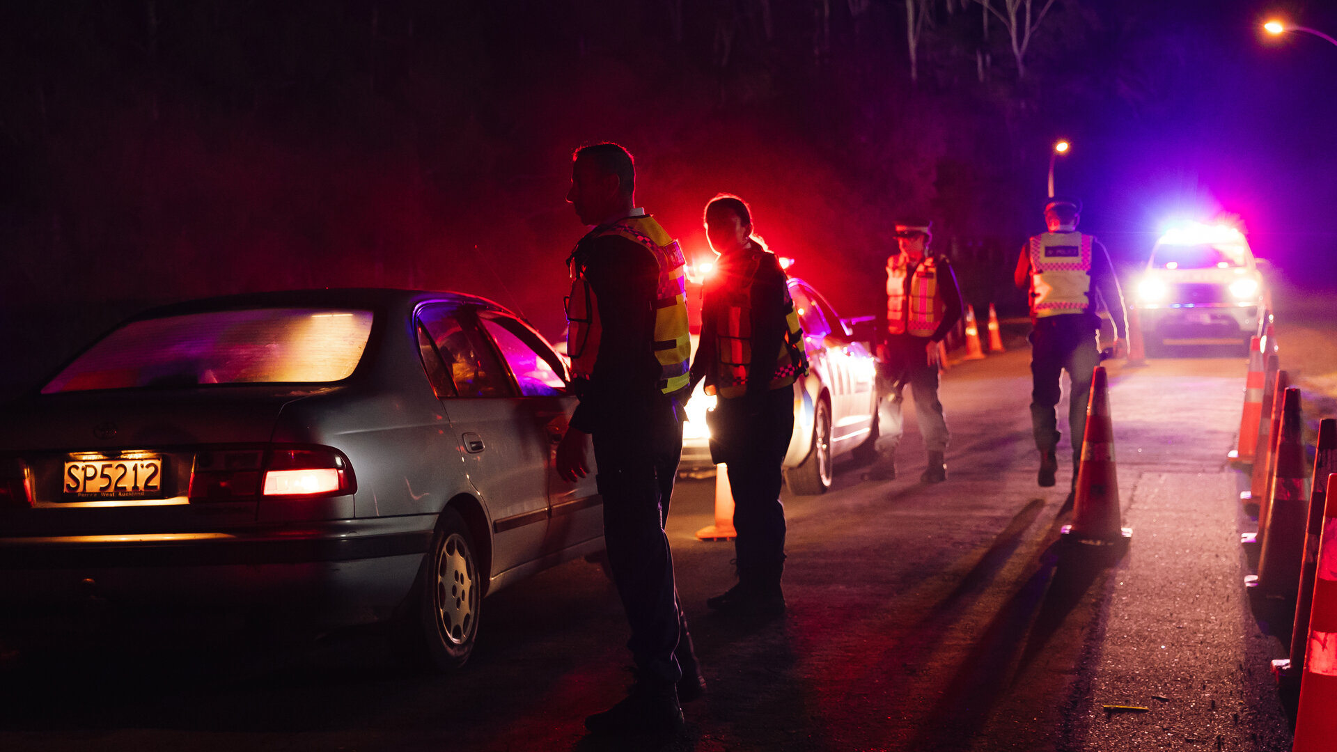 "Back Up - Auf Streife mit der Ex: Das Gesetz der Lieferkette": Bei einer nächtlichen Polizeikontrolle steht ein Auto steht hinter einem Polizeiwagen. Neben dem angehaltenen Fahrtzeug stehen zwei Personen (Komparsen) in dunkler Kleidung und gelben Schutzwesten. Neben dem Polizeiwagen stehen zwei Polizisten (Komparsen) in Polizeiuniformen und gelbe Schutzwesten. Im Hintergrund steht ein größerer Wagen mit eingeschaltetem Licht, welches die Straße ein wenig ausleuchtet.