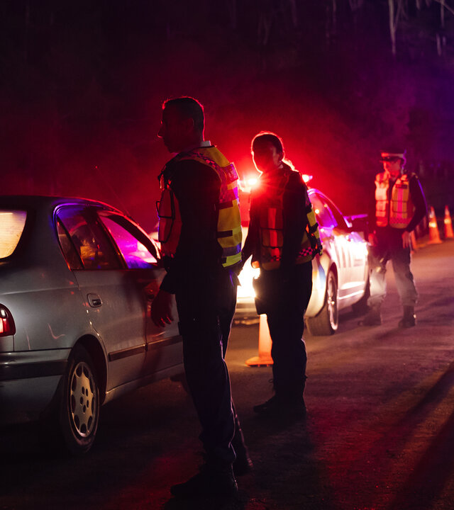 "Back Up - Auf Streife mit der Ex: Das Gesetz der Lieferkette": Bei einer nächtlichen Polizeikontrolle steht ein Auto steht hinter einem Polizeiwagen. Neben dem angehaltenen Fahrtzeug stehen zwei Personen (Komparsen) in dunkler Kleidung und gelben Schutzwesten. Neben dem Polizeiwagen stehen zwei Polizisten (Komparsen) in Polizeiuniformen und gelbe Schutzwesten. Im Hintergrund steht ein größerer Wagen mit eingeschaltetem Licht, welches die Straße ein wenig ausleuchtet.