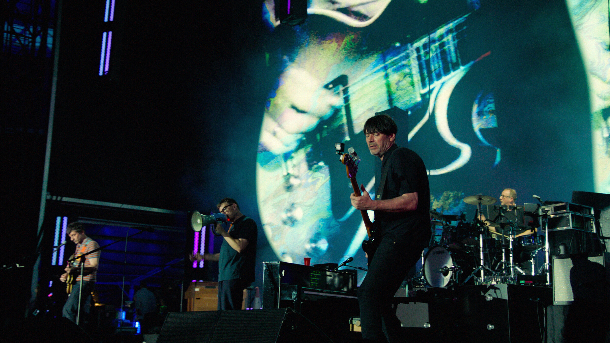"Blur: Live at Wembley Stadium": Die Mitglieder der Band Blur stehen nebeneinander auf einer Bühne mit ihren Instrumenten. Das Geschehen auf der Bühne wird auf eine rückwärtige Videowall projiziert.