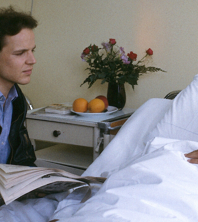 "Diese Drombuschs - Die Machtprobe": Chris (Mick Werup) sitzt am Krankenbett seines Kollegen Wembacher (Claus Derlinghof). Wembacher hat einen Verband am Kopf. Chris hält eine Zeitung in den Händen.