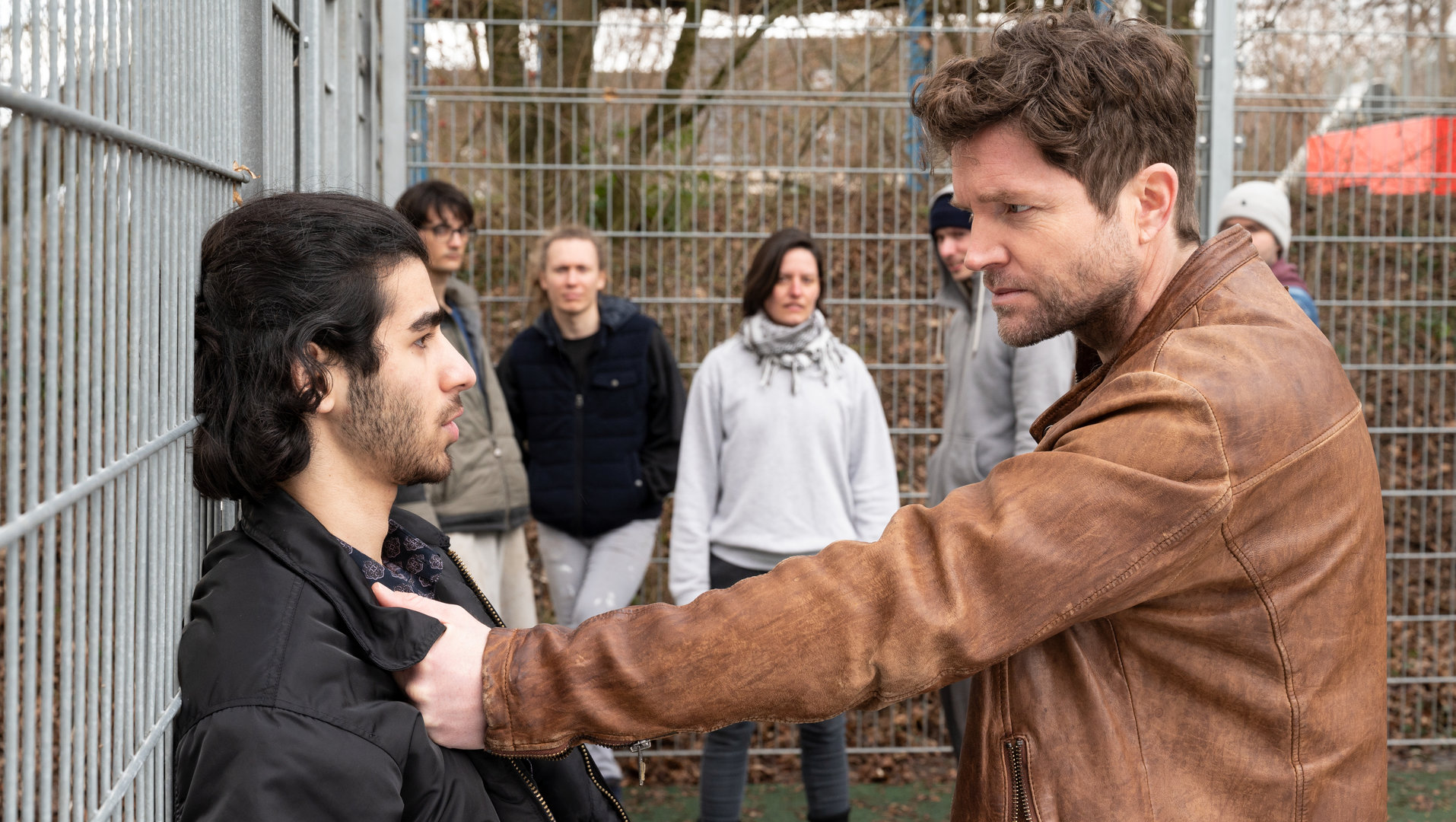 "SOKO Stuttgart – Doppeltes Spiel (1)": Jo Stoll (Peter Ketnath) stellt Kadir Sabia (Doguhan Kabadayi) auf einem Sportplatz zur Rede. Im Hintergrund sammeln sich Schaulustige, während Jo den Verdächtigen am Kragen packt und ihn mit aufforderndem Blick gegen den Zaun drückt.