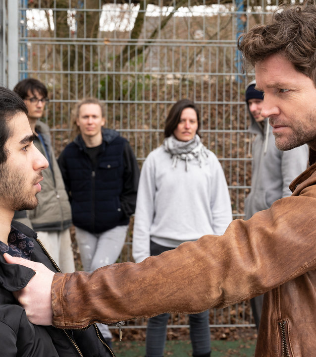 "SOKO Stuttgart – Doppeltes Spiel (1)": Jo Stoll (Peter Ketnath) stellt Kadir Sabia (Doguhan Kabadayi) auf einem Sportplatz zur Rede. Im Hintergrund sammeln sich Schaulustige, während Jo den Verdächtigen am Kragen packt und ihn mit aufforderndem Blick gegen den Zaun drückt.
