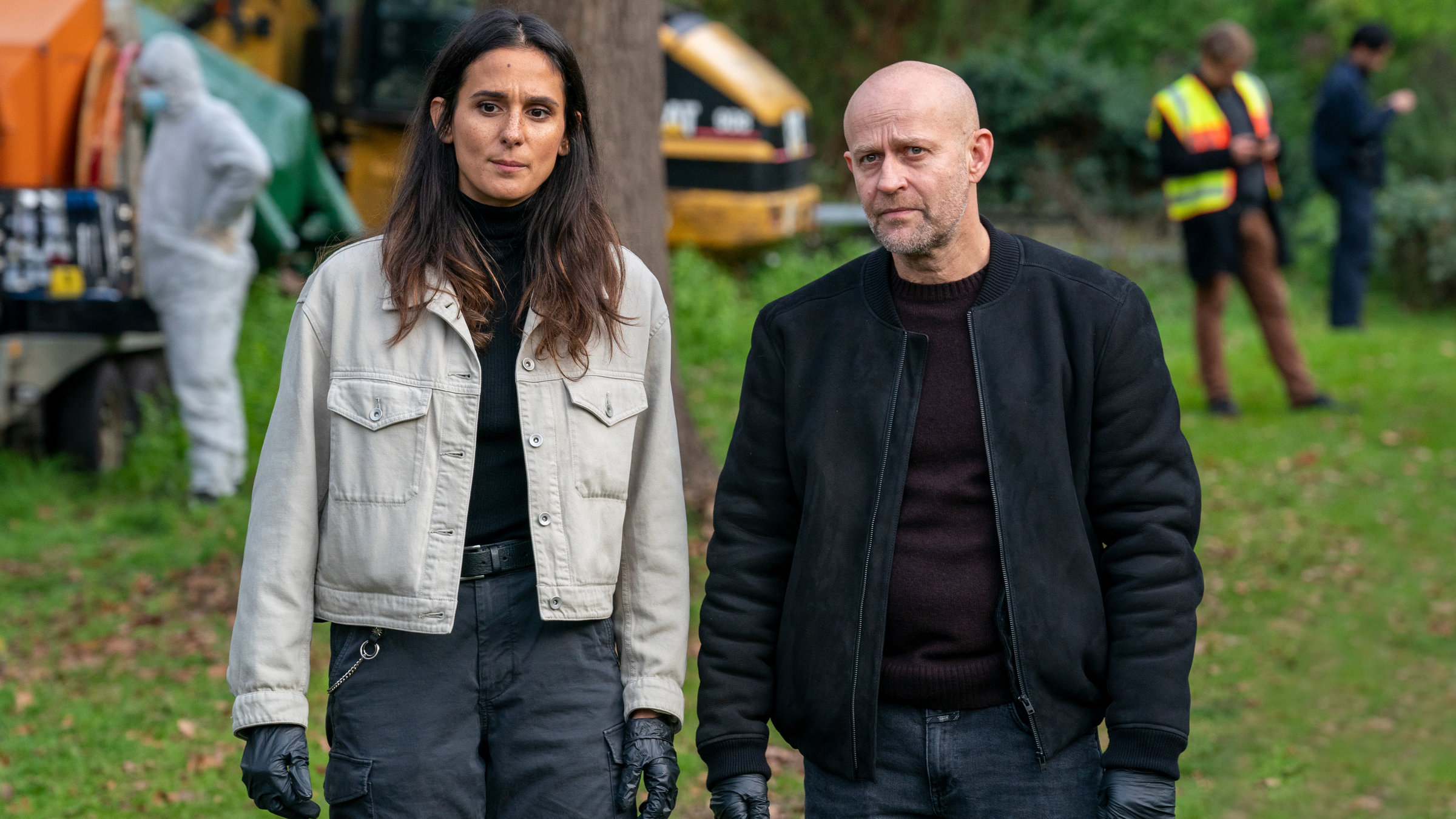 "Jenseits der Spree - Spurlos":  Mavi Neumann (Aybi Era) und Robert Heffler (Jürgen Vogel) stehen auf einer Wiese. Im Hintergrund stehen mehrere Polizisten zwischen Baumaschinen.