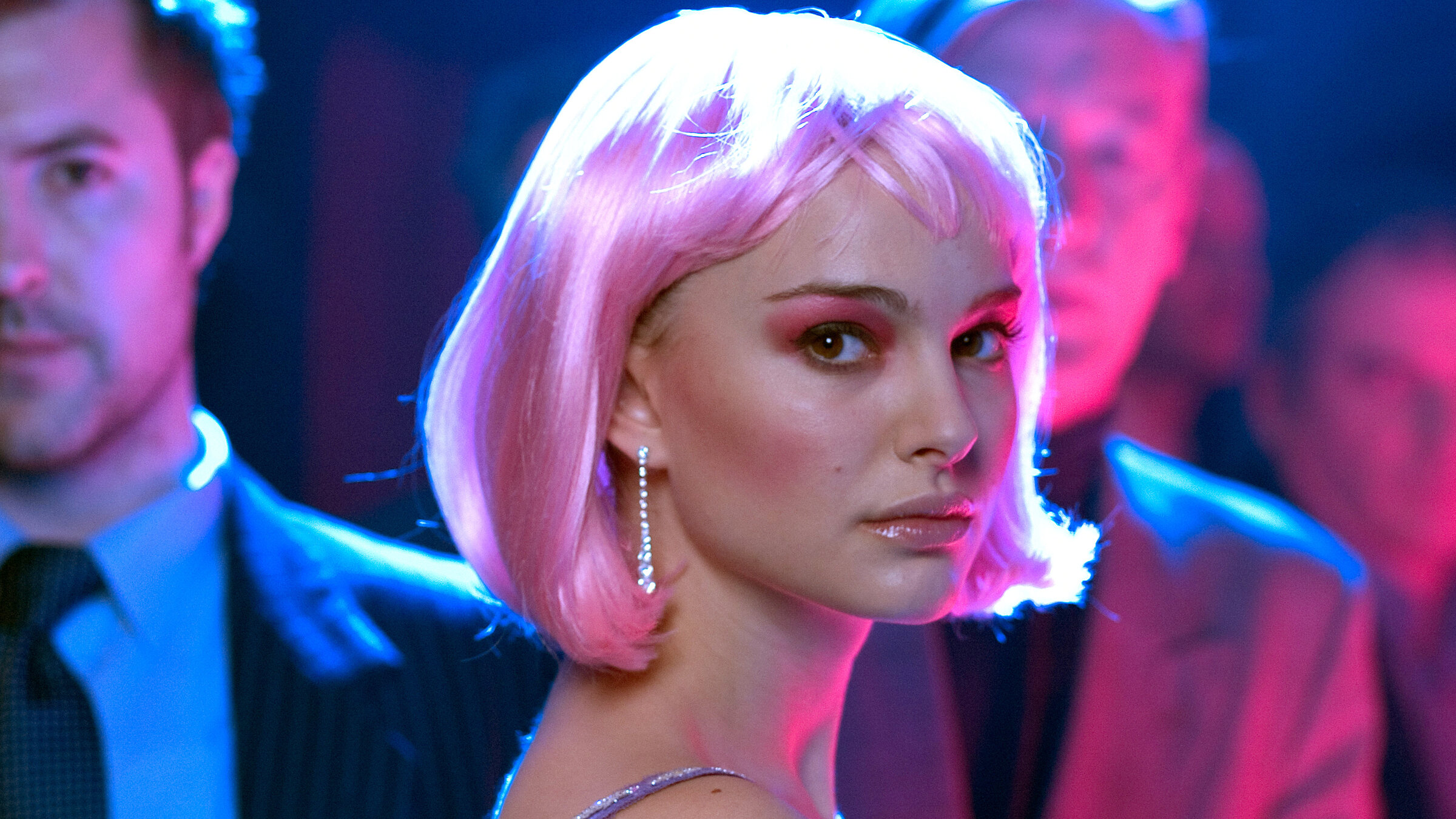 "Hautnah": Alice (Natalie Portman) trägt eine pinkfarbene Perücke, auffälliges Make-up und blickt ernst in die Kamera. Sie steht inmitten einer Gruppe von Menschen in einem in blaues und pinkes Licht getauchten Raum.