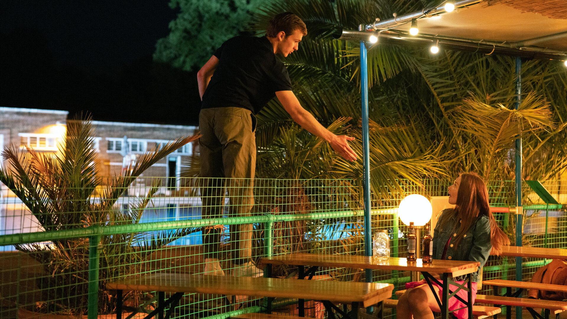 "You & Me": Ben (Harry Lawtey) steht auf der anderen Seite des Zauns und reicht Emma (Jessica Barden), die in einem Biergarten sitzt und ihn überrascht ansieht, die Hand.