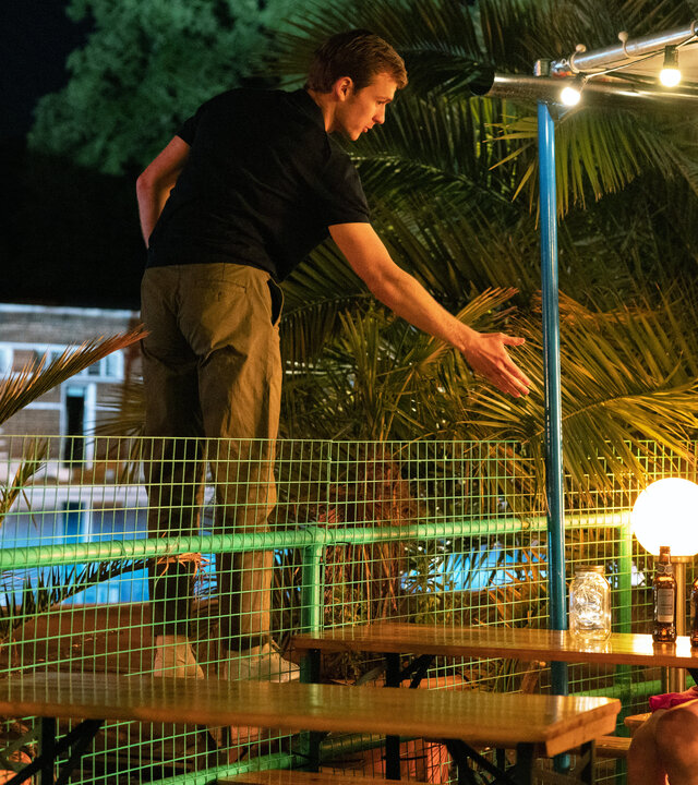 "You & Me": Ben (Harry Lawtey) steht auf der anderen Seite des Zauns und reicht Emma (Jessica Barden), die in einem Biergarten sitzt und ihn überrascht ansieht, die Hand.