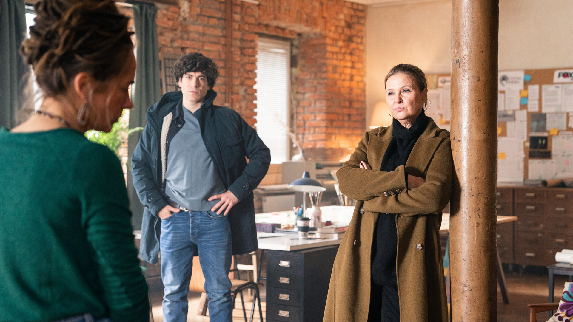 "Die Chefin - Mittel zum Zweck": In einem Büroraum: Korbinian Kirchner (Jonathan Hutter) hat die Hände in die Hüften gestemmt. Vera Lanz (Katharina Böhm) hat die Arme vor der Brust verschränkt und lehnt gegen eine Säule im Raum. Ihnen gegenüber steht Sophie Albrecht (Annina Hellenthal). Sie weicht dem Blick der Ermittler aus. Im Hintergrund stehen Schreibtische vor einer Wand aus Backstein. An einer weiteren Wand hängen Pinnwände mit ausgedruckten Notizblättern und Sticky Notes.