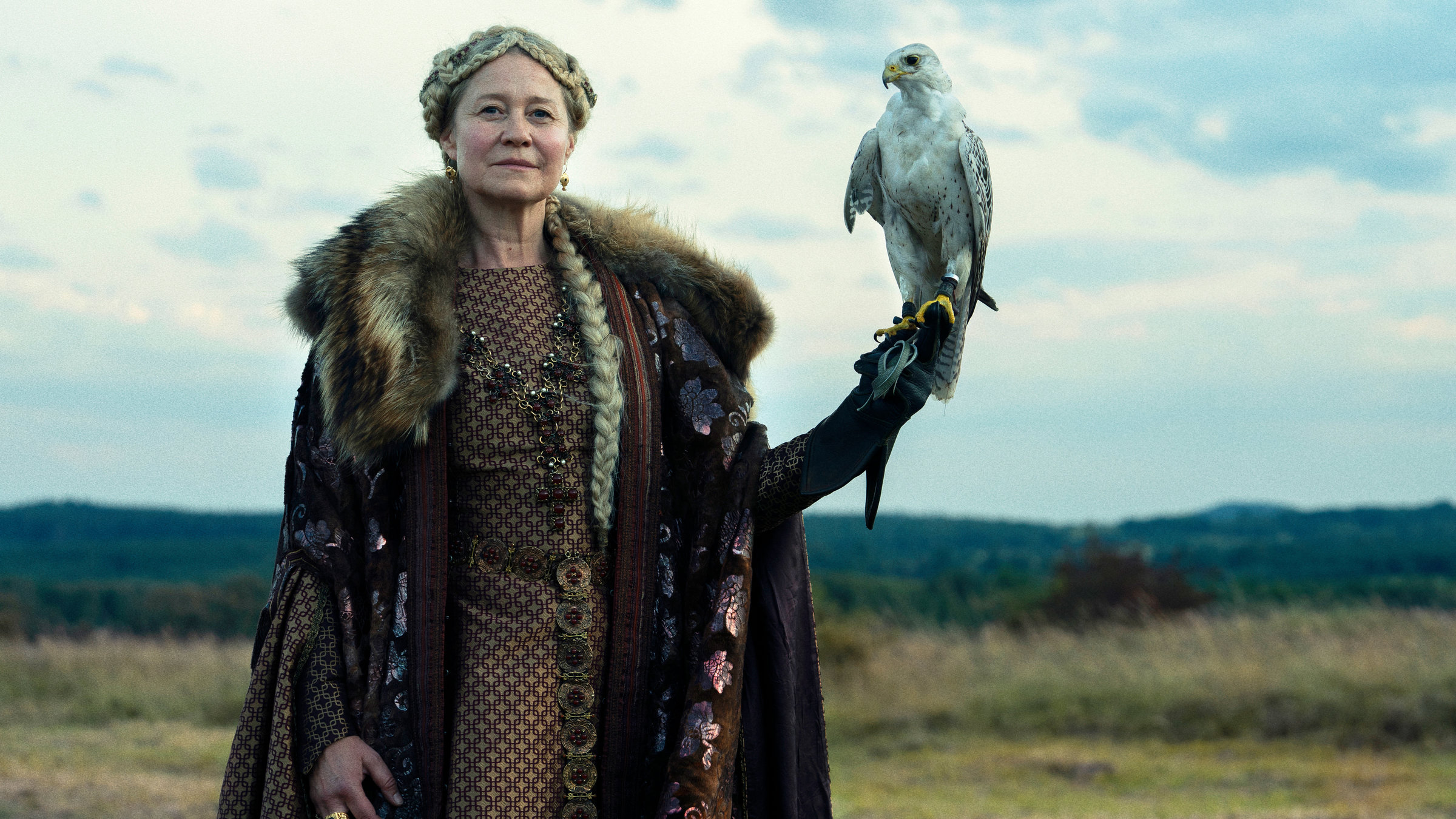 "Die Königin des Nordens": Prunkvoll in einem prächtigen Kleid und Mantel mit Pelzkragen demonstriert Margarethe I. (Trine Dyrholm) mit einem Falken auf der ausgestreckten Hand ihre Macht. Sie steht in einer weiten, flachen Landschaft.