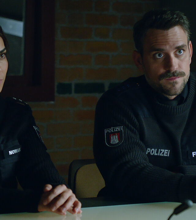"Notruf Hafenkante - Helfer in Not": Die Polizistin Isa Nowak (Lilli Hollunder) und ihr Kollege Kris Freiberg (Marc Barthel) sitzen nebeneinander an einem Tisch im Befragungsraum des PK. Beide sehen in die gleiche Richtung.