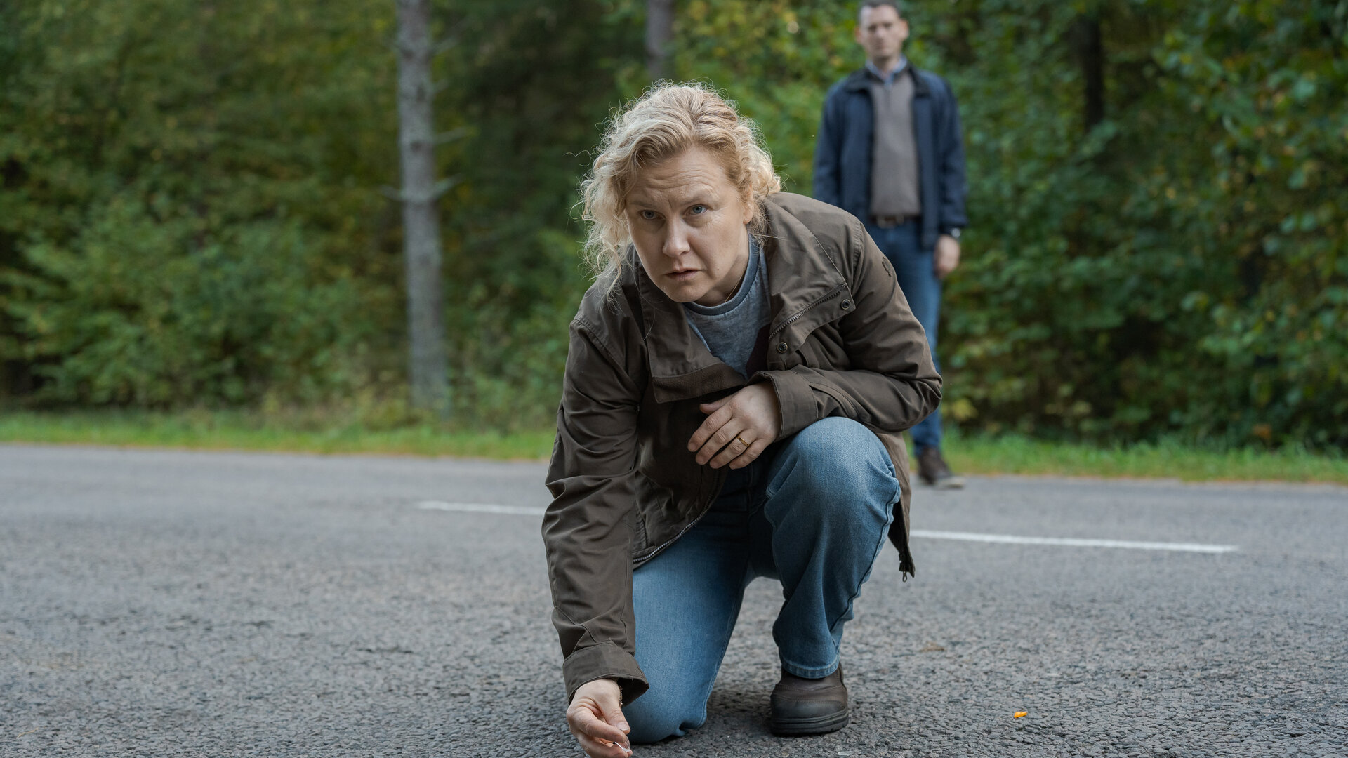 "Wolfssommer – Blutige Spuren - Die Spur der Wölfe": Hannah (Eva Melander) kniet auf einer Straße. Sie hebt etwas mit zwei Fingern auf. Vor ihr auf der Fahrbahn sind kleine Gegenstände zu sehen. Im Hintergrund steht Gordon (Hannes Fohlin) vor einem Waldstück.
