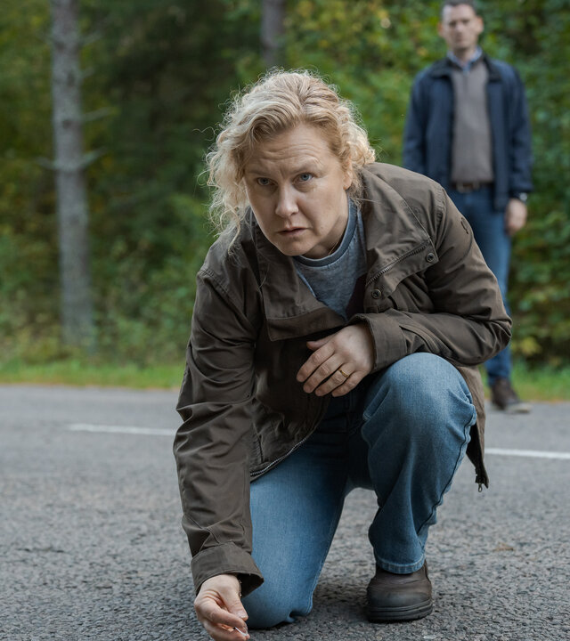 "Wolfssommer – Blutige Spuren - Die Spur der Wölfe": Hannah (Eva Melander) kniet auf einer Straße. Sie hebt etwas mit zwei Fingern auf. Vor ihr auf der Fahrbahn sind kleine Gegenstände zu sehen. Im Hintergrund steht Gordon (Hannes Fohlin) vor einem Waldstück.