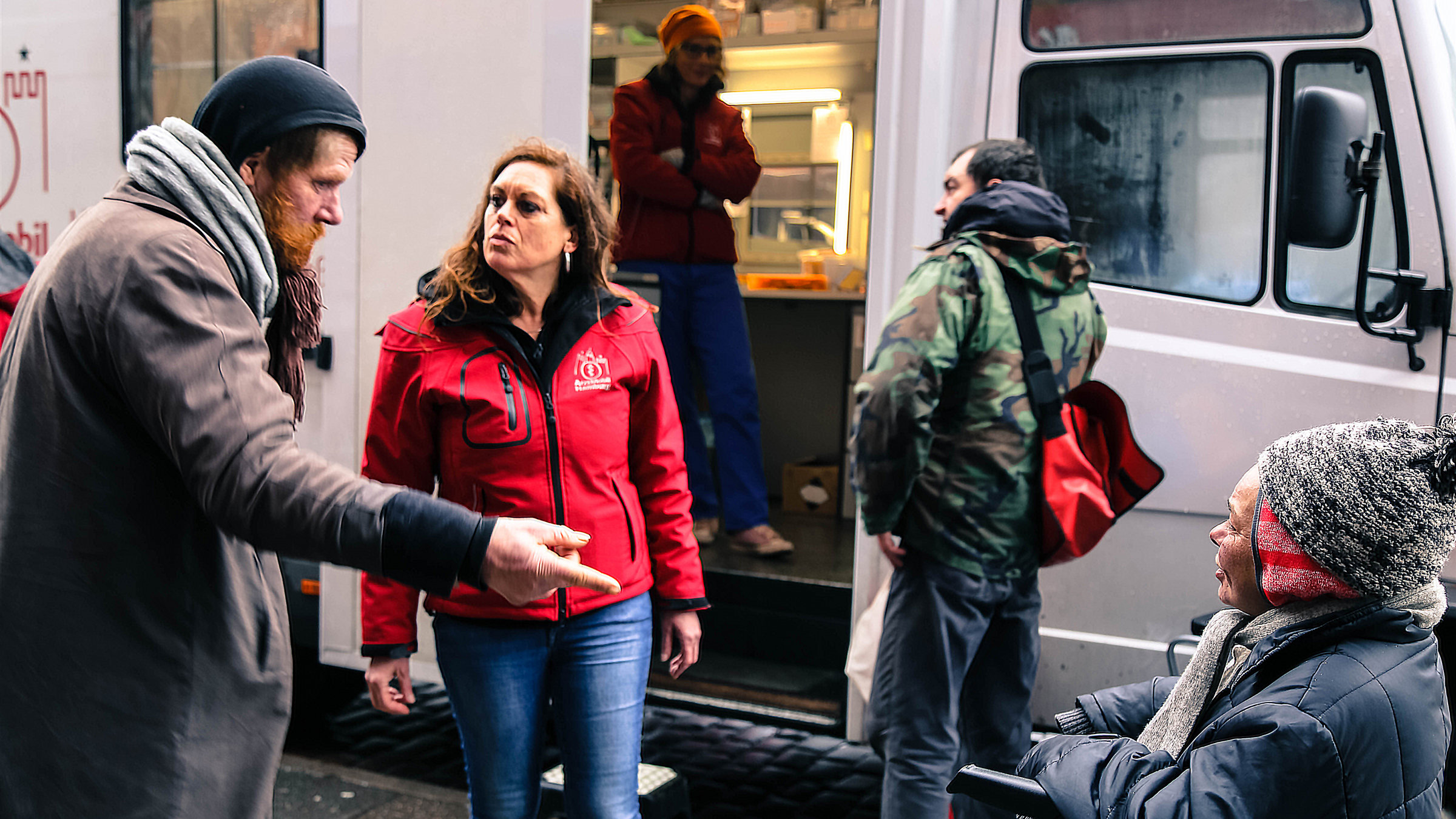 "37°: Wo andere wegsehen - Ärzte im Einsatz für Obdachlose": Levke Sonntag steht vor dem Arztmobil und schaut auf einen Obdachlosen. Rechts im Bild vor dem Bus stehen zwei weitere Obdachlose.