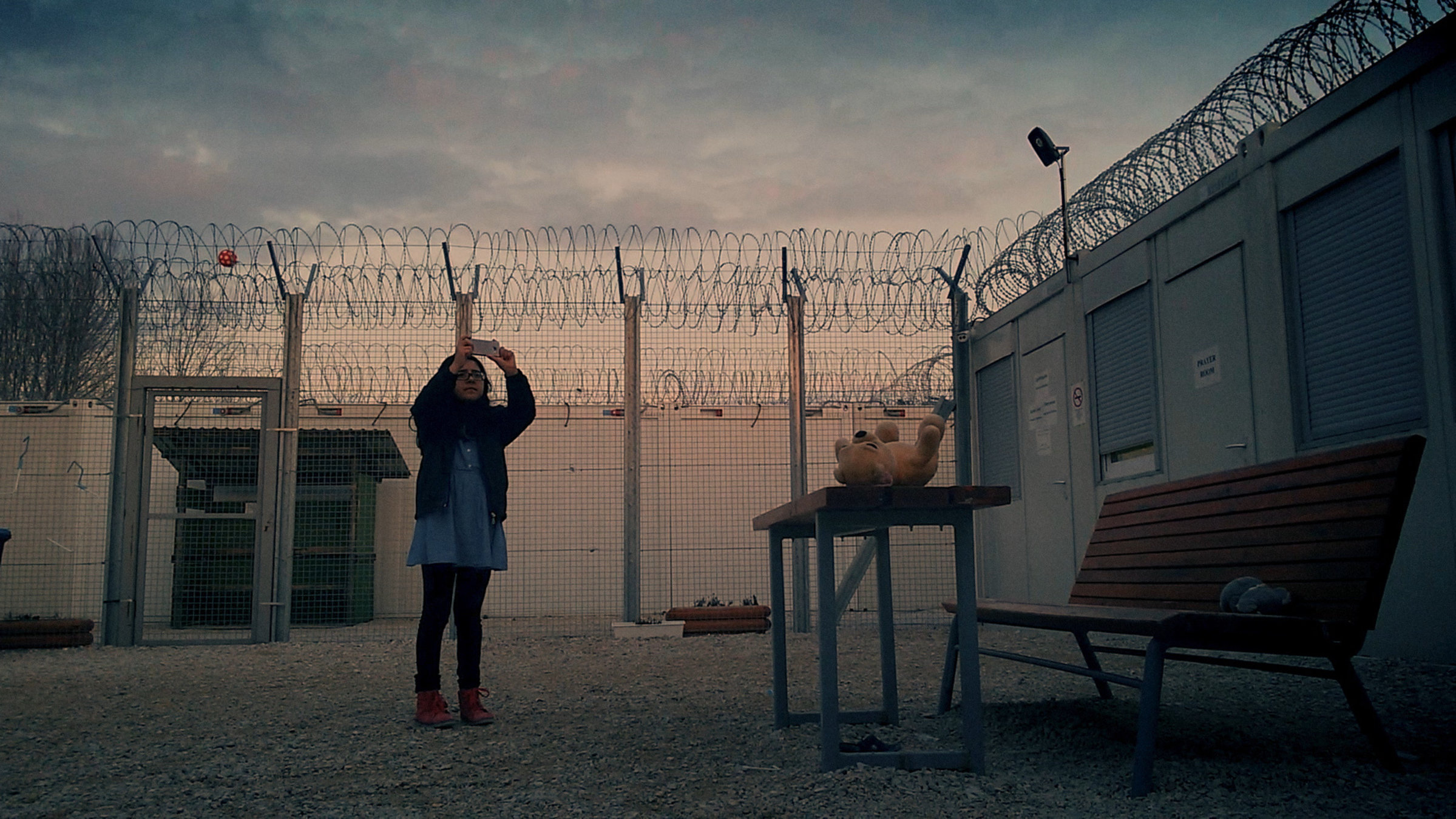 "Midnight Traveller": Mädchen filmt mit Handy in einem mit Stacheldraht abgezäunten Hof. Ihr Teddybär liegt derweil auf einem kleinen Tisch im Vordergrund rechts.