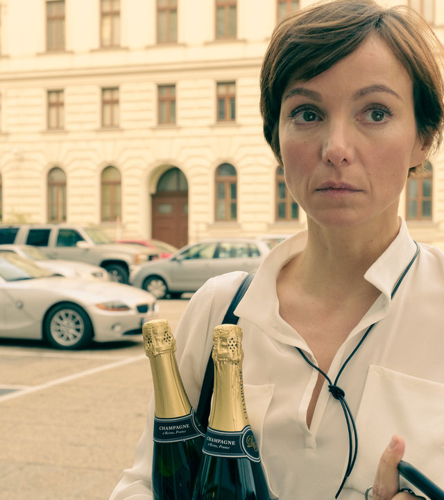 "Am Anschlag - Die Macht der Kränkung - MIRA": Mira (Julia Koschitz) blickt in die Ferne. In der linken Hand hält sie zwei Champagner Flaschen und in der rechten Hand ihr Mobiltelefon und einen Schlüsselbund. Im Hintergrund ist ein Gebäude zu sehen, vor dem Autos parken.