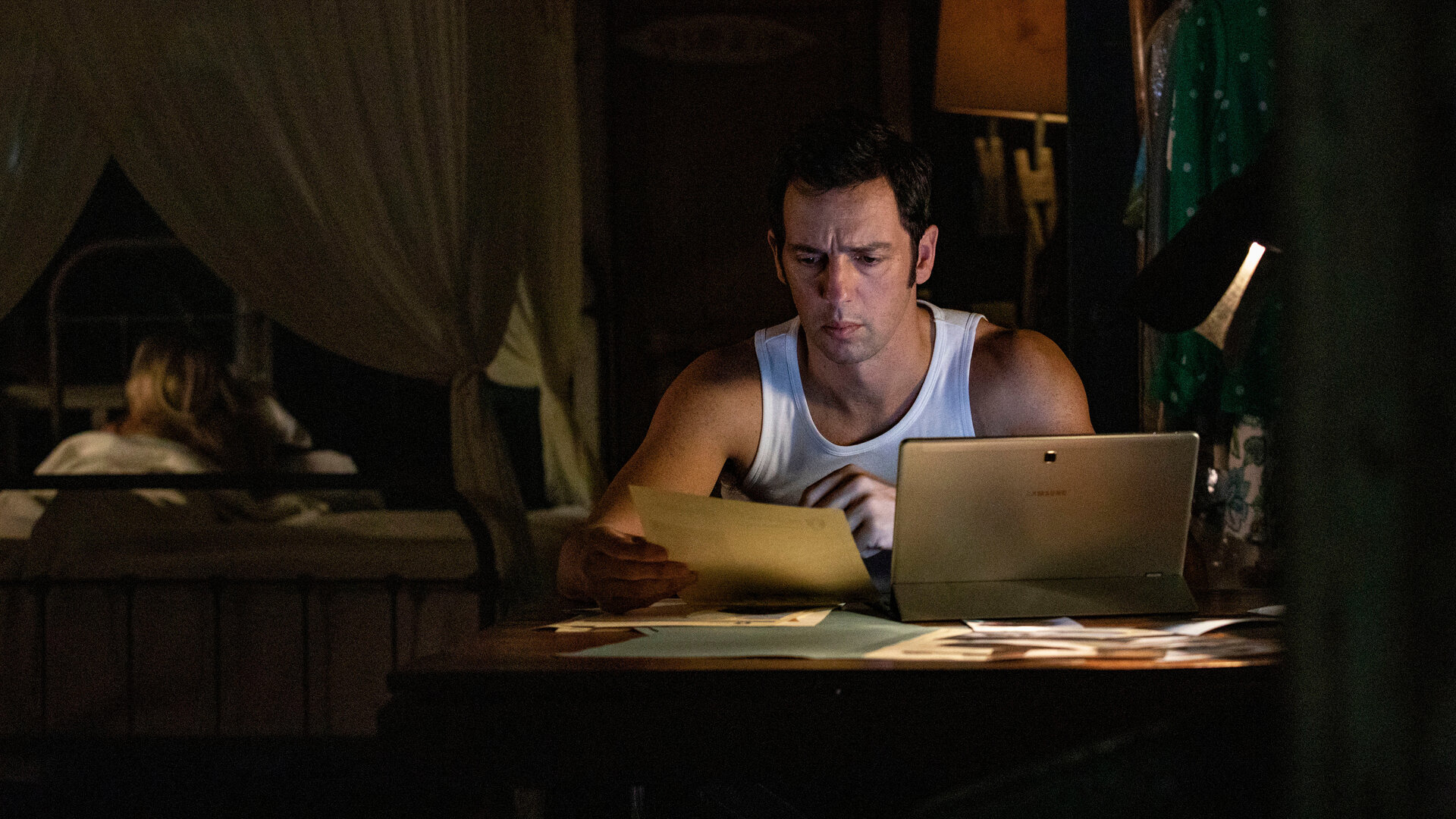 "Death in Paradise - Das Spiel": Detective Neville Parker (Ralf Little) sitzt zu Hause im Dunkeln vor seinem Laptop. In der Hand hält er ein Dokument, das er akribisch liest.