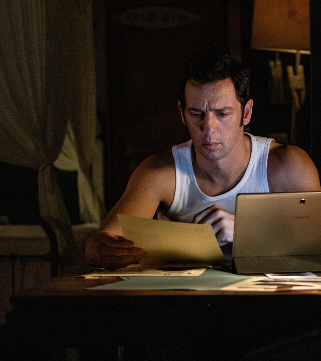 "Death in Paradise - Das Spiel": Detective Neville Parker (Ralf Little) sitzt zu Hause im Dunkeln vor seinem Laptop. In der Hand hält er ein Dokument, das er akribisch liest.