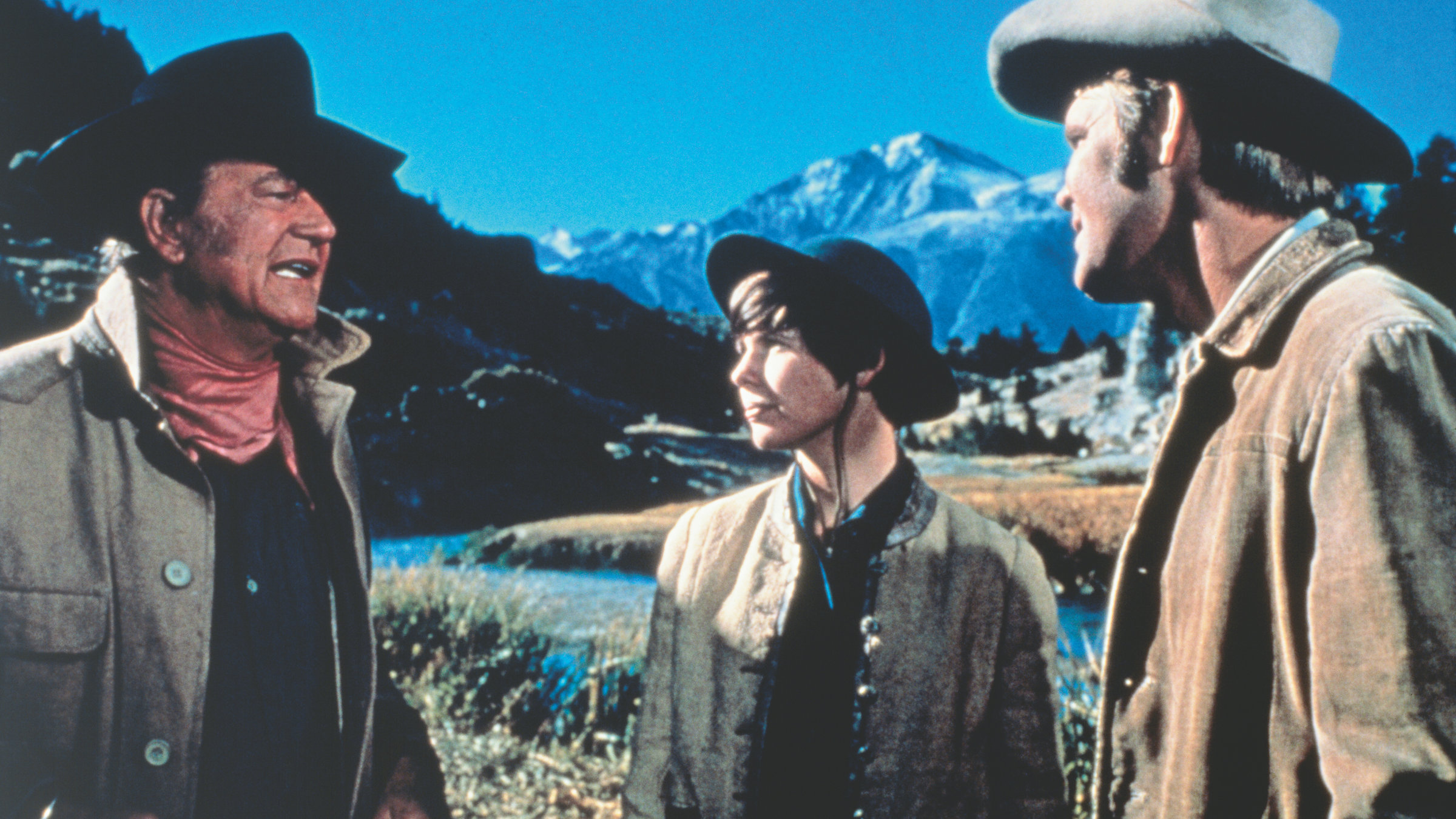 "Der Marshal": ": Ein pittoreskes felsiges Tal mit einem geschlängelten Flusslauf im Vordergrund und einem schneebedeckten Bergmassiv am Horizont. Rooster Cogburn (John Wayne), Mattie Ross (Kim Darby) und La Boeuf (Glen Campbell) stehen am Flussufer zusammen; Cogburn spricht, die beiden anderen schauen zu ihm.