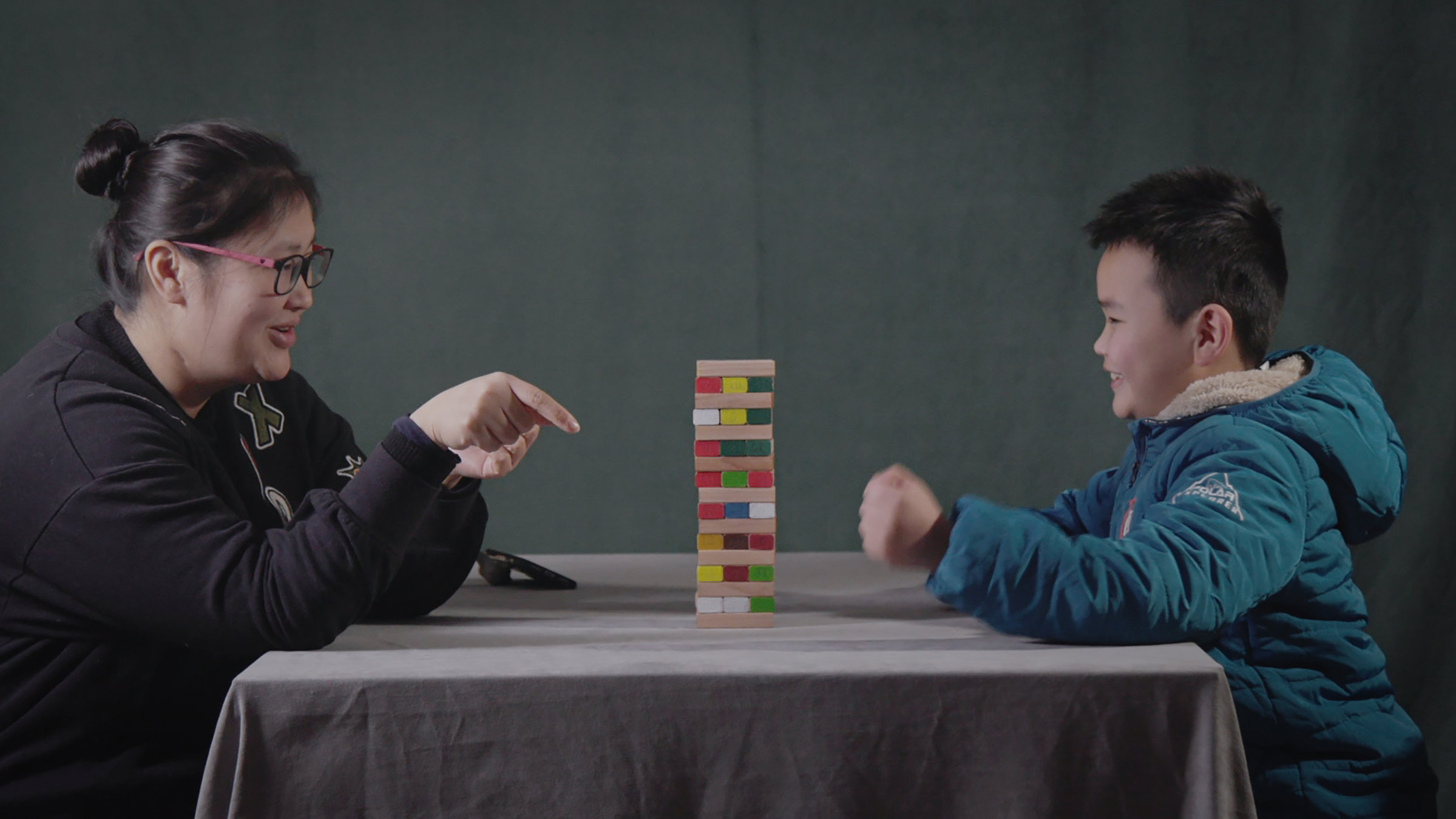 "Glücklich bin ich, wenn Du schläfst": Auf einem Tisch steht ein Spiel aus Holzklötzchen. Die Mutter Qi Jingjing und ihr Sohn Liu Yufeng sitzen sich an dem Tisch gegenüber und spielen das Spiel Jenga mit den Holzklötzen. Beide haben Spaß dabei.