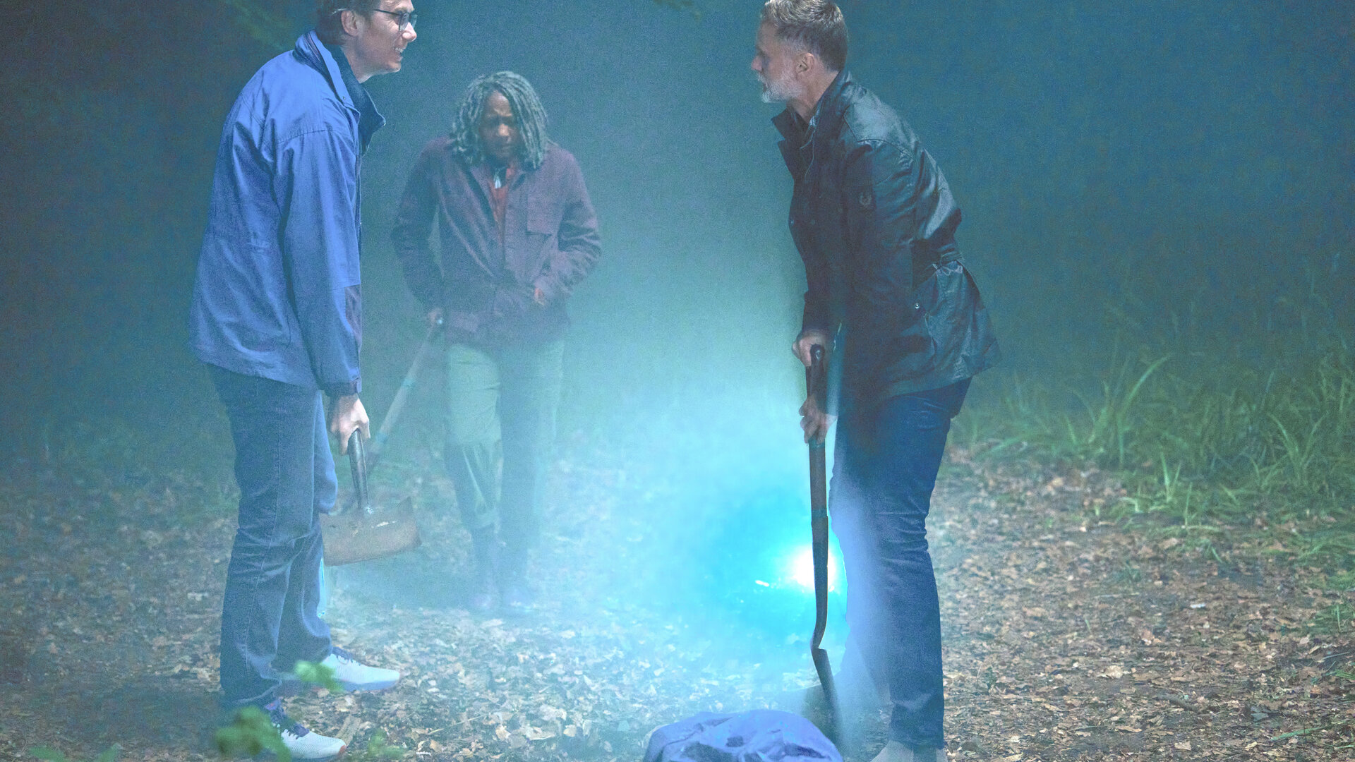 "The Outlaws - Bullenauge": Greg (Stephen Merchant), Myrna (Clare Perkins) und John (Darren Boyd) stehen auf einer mit einer Taschenlampe beleuchteten Lichtung und haben Spaten in der Hand. Vor ihnen liegt ein Sack, in den etwas gehüllt ist.