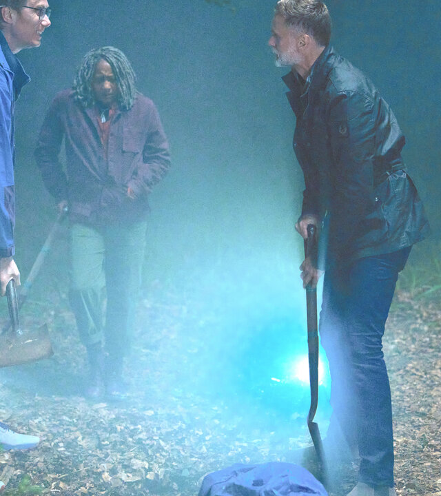 "The Outlaws - Bullenauge": Greg (Stephen Merchant), Myrna (Clare Perkins) und John (Darren Boyd) stehen auf einer mit einer Taschenlampe beleuchteten Lichtung und haben Spaten in der Hand. Vor ihnen liegt ein Sack, in den etwas gehüllt ist.