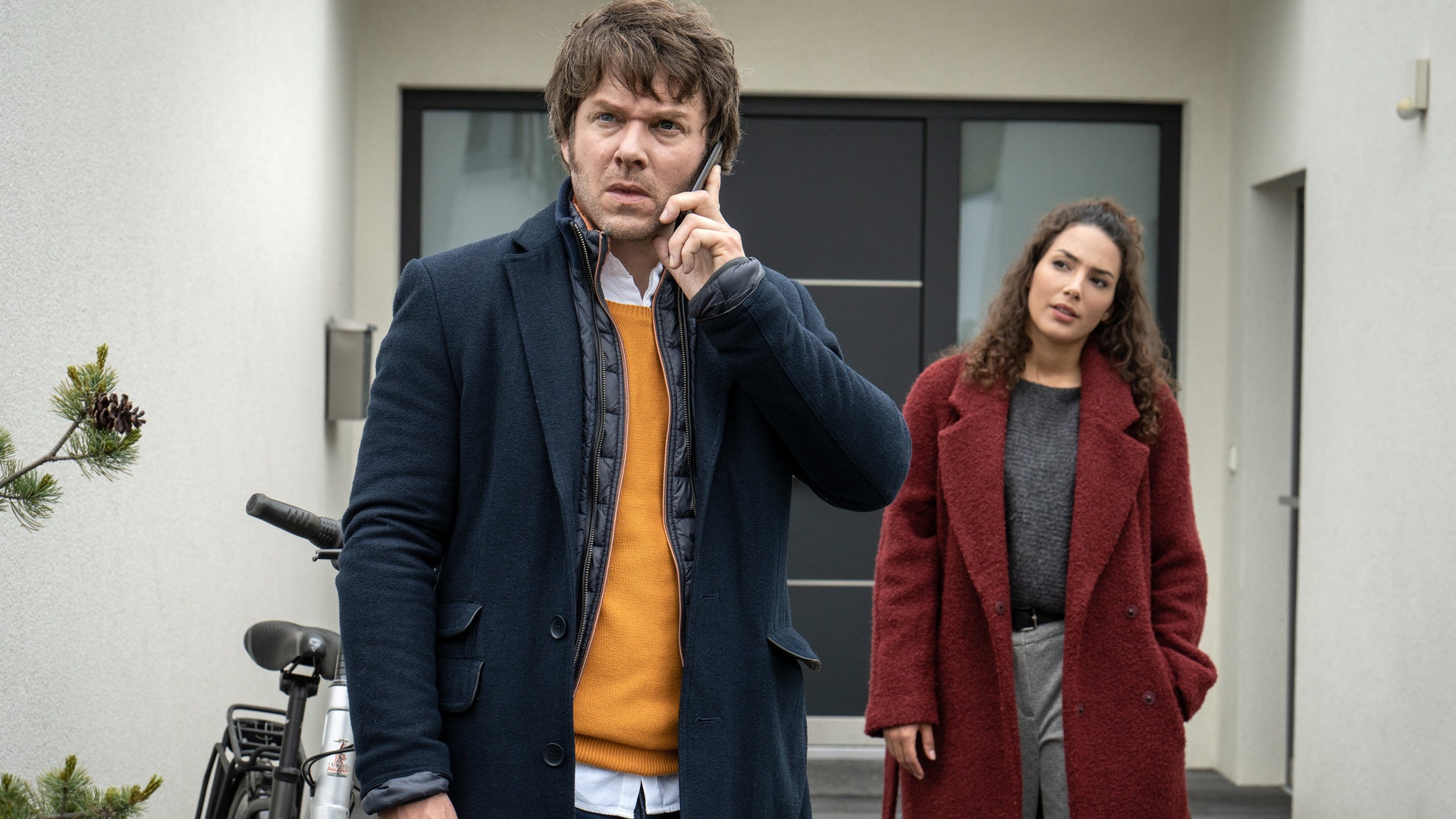 "SOKO Leipzig - Kowalskis Entscheidung": Tom Kowalski (Steffen Schroeder) steht gemeinsam mit Kim Nowak (Amy Mußul) vor einem Hauseingang. Er steht mit dem Rücken zu Kim und telefoniert. Sie schaut ihn verständnisvoll an.