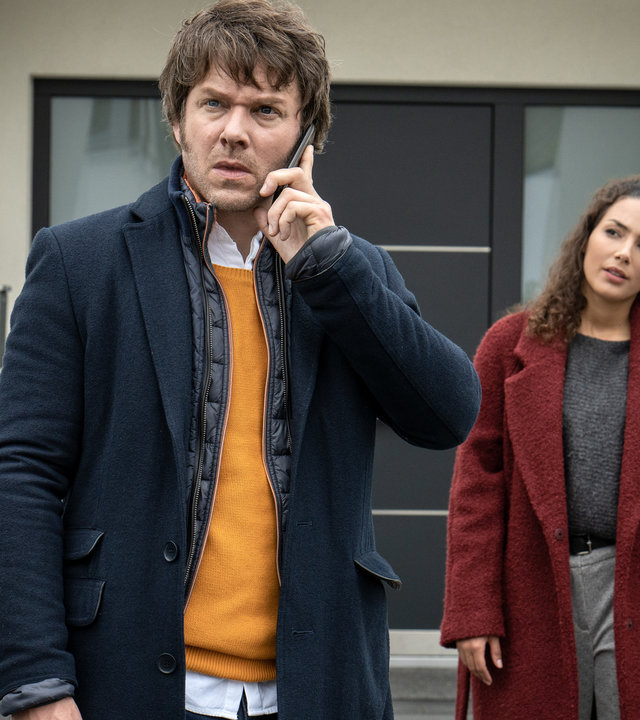 "SOKO Leipzig - Kowalskis Entscheidung": Tom Kowalski (Steffen Schroeder) steht gemeinsam mit Kim Nowak (Amy Mußul) vor einem Hauseingang. Er steht mit dem Rücken zu Kim und telefoniert. Sie schaut ihn verständnisvoll an.