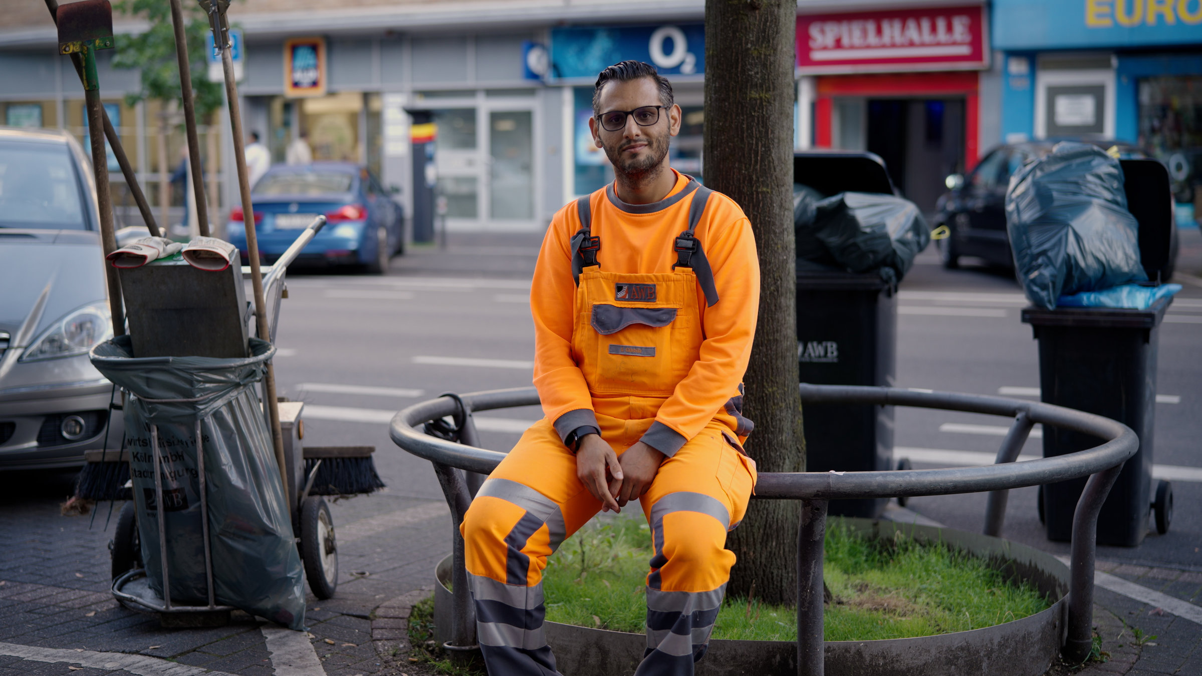 "ZDF.reportage: Müll an jeder Ecke - Stadtreinigung am Limit": Serkan sitzt in orangefarbener Arbeitskleidung auf einem Geländer an der Straße. Neben ihm steht sein Kehrwagen.