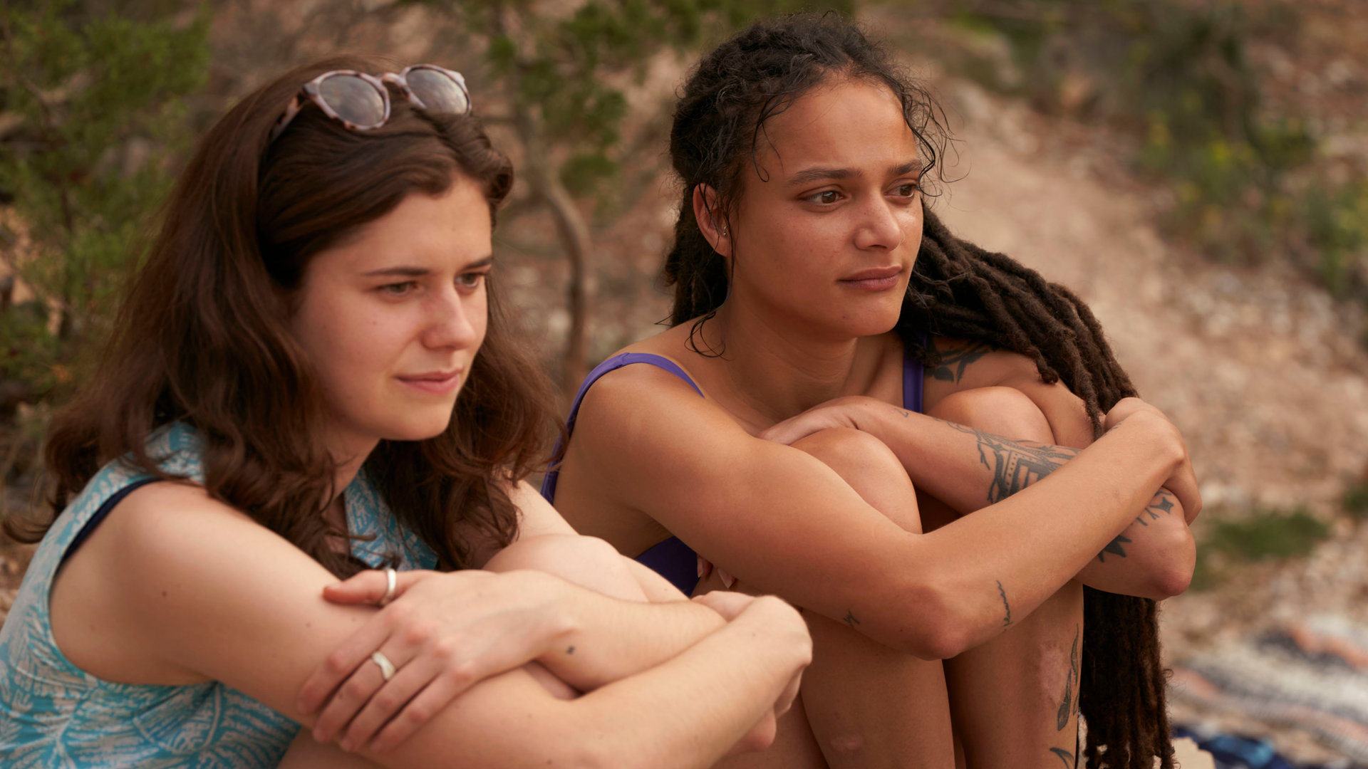 "Conversations with Friends - Der Urlaub": Frances (Alison Oliver) und Bobbi (Sasha Lane) sitzen nebeneinander zusammengekauert am Strand und schauen gedankenvertieft in die Ferne.