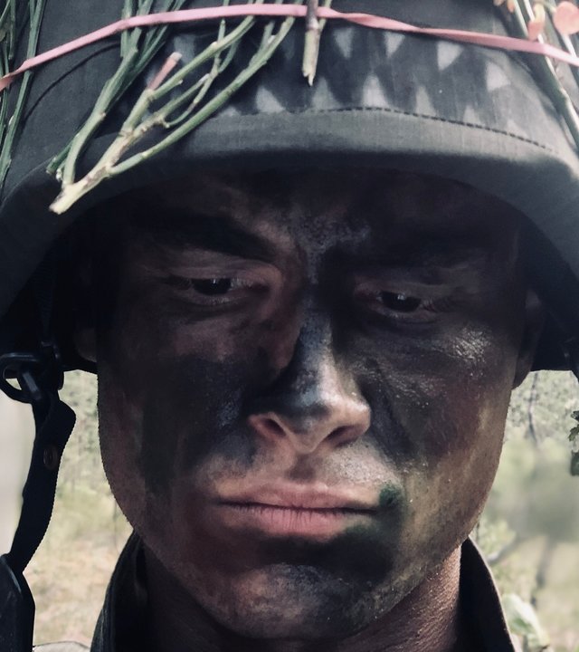 "Californian Commando - Camping": Van (Kian Lawley) steht mit dunkler Gesichtsbemalung und Ästen zur Tarnung im Wald und schaut bedrückt nach unten.