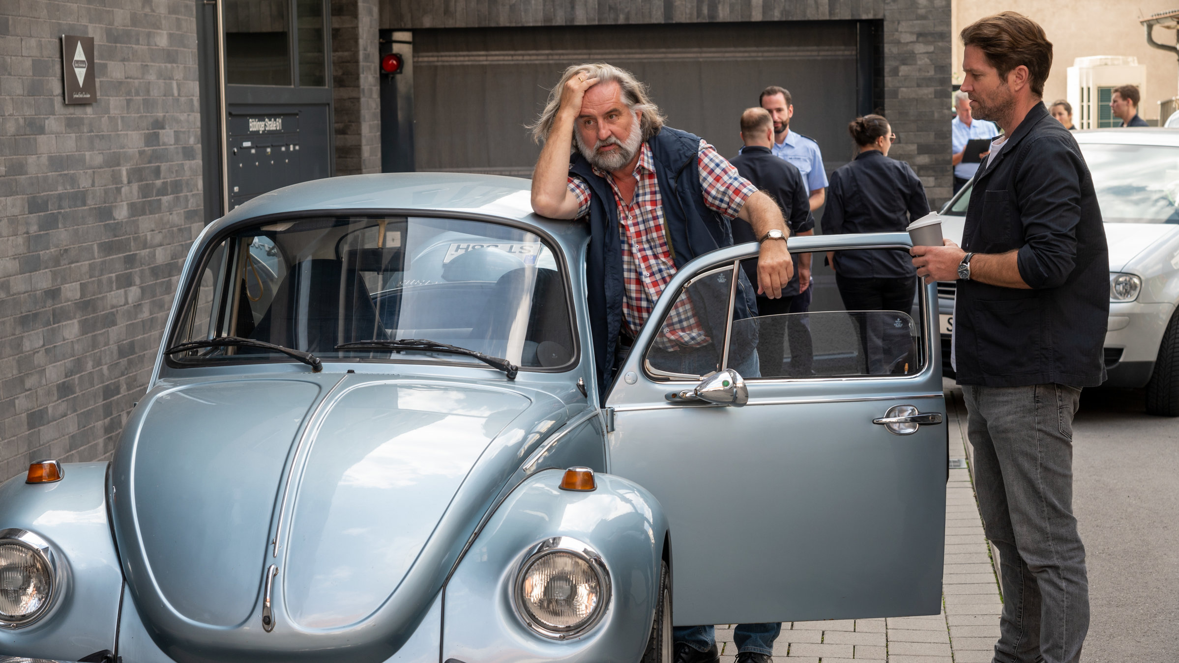 "SOKO Stuttgart - Süßer Tod": Kriminalkommissar Jo Stoll (Peter Ketnath) steht an der offenen Tür eines hellblauen VW Käfers. Zwischen Fahrertür und Auto steht Bernhard "Balu" Völk (Andreas Windhuis), ein Mitarbeiter der Schokoladenfabrik "Schoko Seitz". Balu hält sich die Hand an seine Stirn, während er bedrückt in die Ferne schaut. Im Hintergrund sieht man weitere Streifenpolizisten.