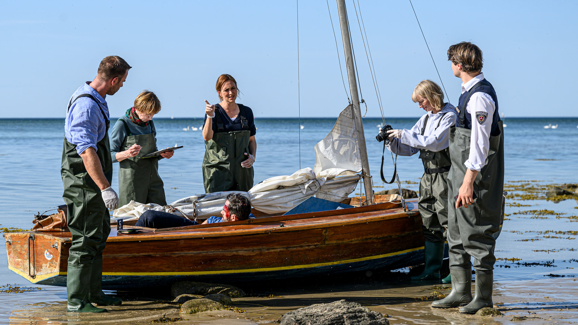 "SOKO Wismar – Tod und Korn": Am Strand, in einem Segelboot findet das Team Peter Braun (Thomas Welsand) tot vor. Im flachen Wasser um das Boot herum stehen Lars Pöhlmann (Dominic Boeer), Karoline Joost (Nike Fuhrmann), Helene Sturbeck (Katharina Blaschke), Roswitha Prinzler (Silke Matthias), Eddi Jansons (Gustavs Gailus).