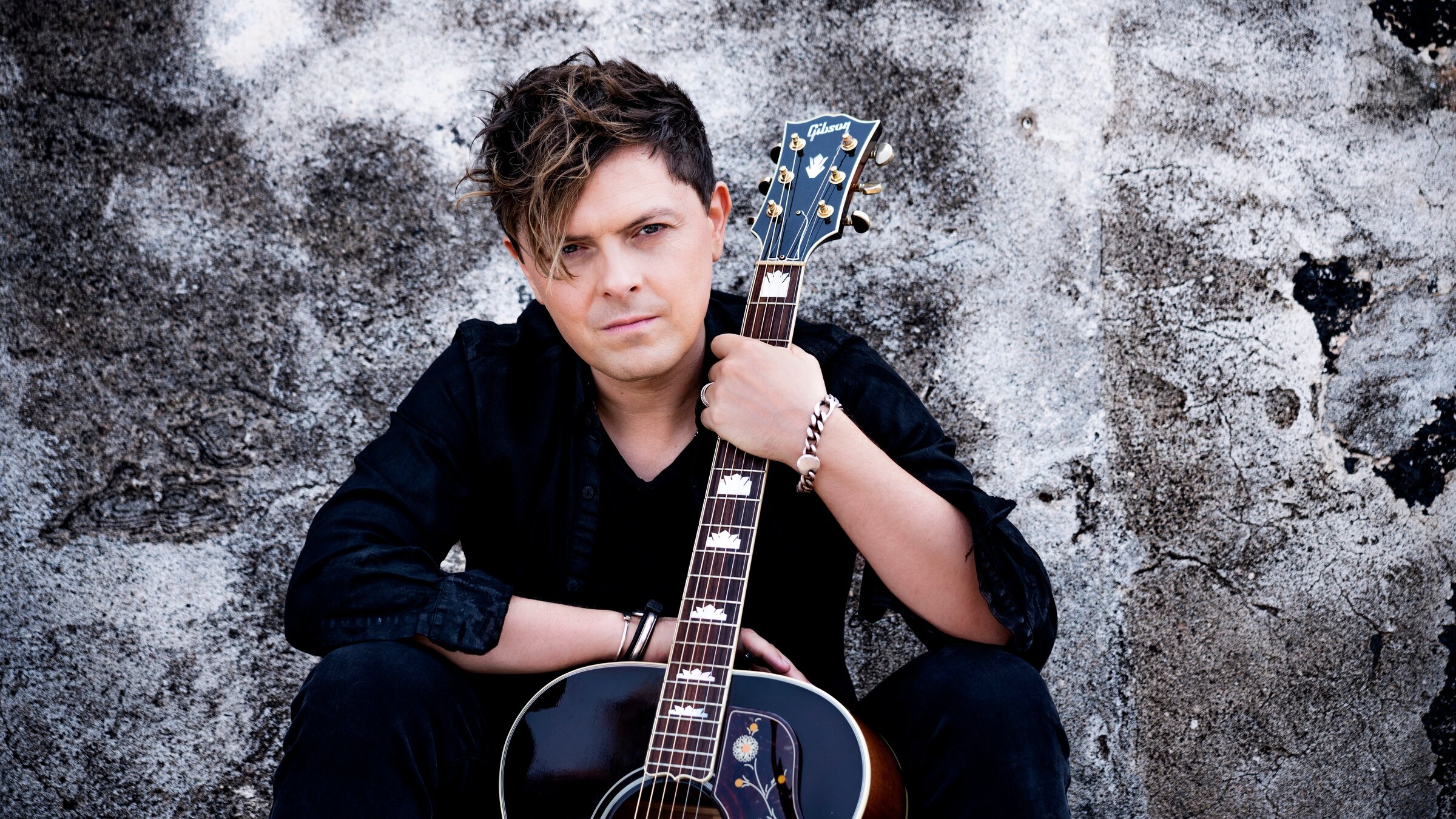 "Michael Patrick Kelly: B.O.A.T.S. - LIVE": Der Musiker Michael Patrick Kelly sitzt vor einer grauen Wand. Er blickt in die Kamera. Vor ihm auf dem Boden steht eine Gitarre, die er mit der linken Hand festhält. Gekleidet ist er mit schwarzer Hose und schwarzem Hemd.