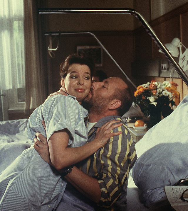 "Die Schwarzwaldklinik - Die Rentnerinitiative": Der Patient Körtler (Klaus Guth) liegt im Bett und zieht Schwester Ina (Gaby Fischer) zu sich. Dabei gibt er ihr einen Kuss auf die Wange.