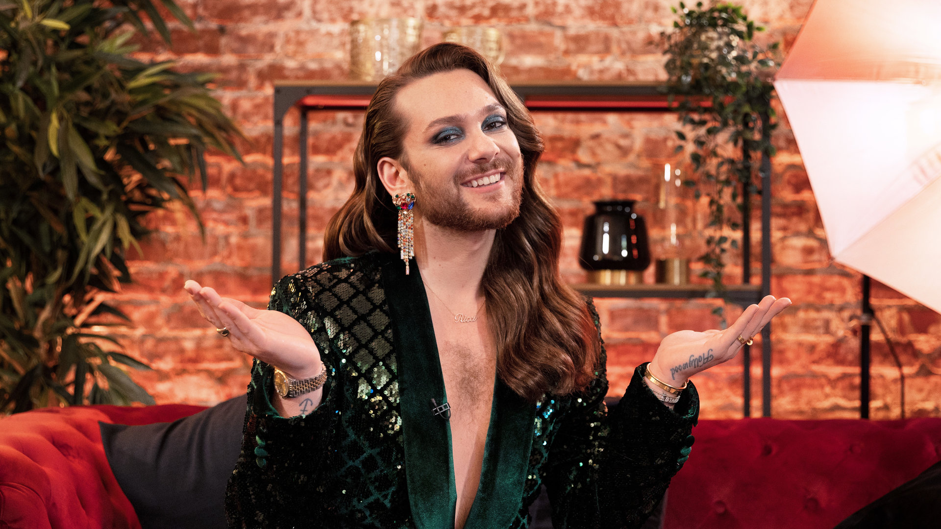 "Glow Up - Deutschlands nächster Make-up-Star: Finale: Kampagne": Riccardo Simonetti sitzt auf einem roten Sofa in der "Glow Up"-Lounge. Er trägt einen grünen Anzug und lächelt in die Kamera.