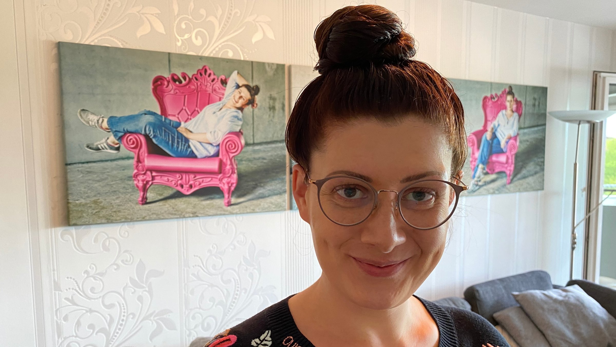 "37° Ungeküsst - Wenn die Liebe auf sich warten lässt": Jana Crämer mit hochgestecktem Haar steht rechts im Bild in einem Wohnzimmer. Sie schaut freundlich in die Kamera. Auf der Wand im Hintergrund hängt ein Foto von ihr, auf dem sie in einem pinken Sessel sitzt und lacht.