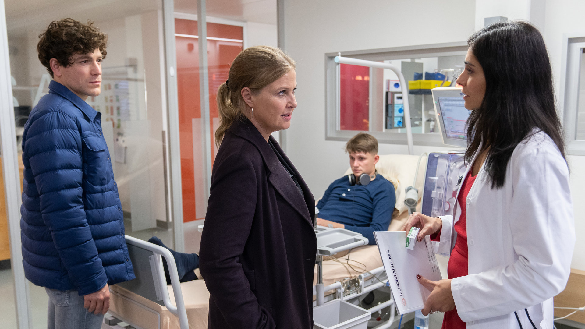 "Die Chefin - Deadline": Das Gespräch im Krankenhaus. Vera Lanz (Katharina Böhm) und Korbinian Kirchner (Jonathan Hutter) stehen der Ärztin Dr. Leyla Sahin (Pegah Ferydoni) gegenüber, sie sind alle von der Seite zu sehen. Im Hintergrund ist das Krankenbett mit dem Patienten Florian Blanke (Leon Blaschke) zu sehen.