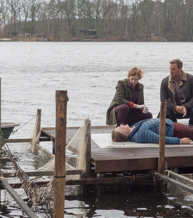 „SOKO Wismar – Kalt serviert“: Auf einem Steg am See liegt die Leiche von Arne Bahlbeck (Klaus Christian Schreiber). Neben ihm knien Helene Sturbeck (Katharina Blaschke) und Lars Pöhlmann (Dominic Boeer). Roswitha Prinzler (Silke Matthias) von der Kriminaltechnik untersucht derweil ein Boot, das auf dem See treibt.